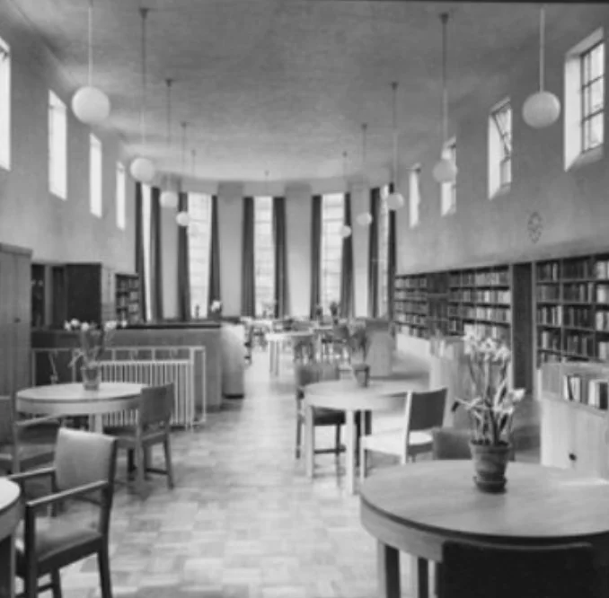 Black and white photograph of the inside of Belsize Library around the time it opened, with tall windows, globe lights, round tables and wooden bookcases